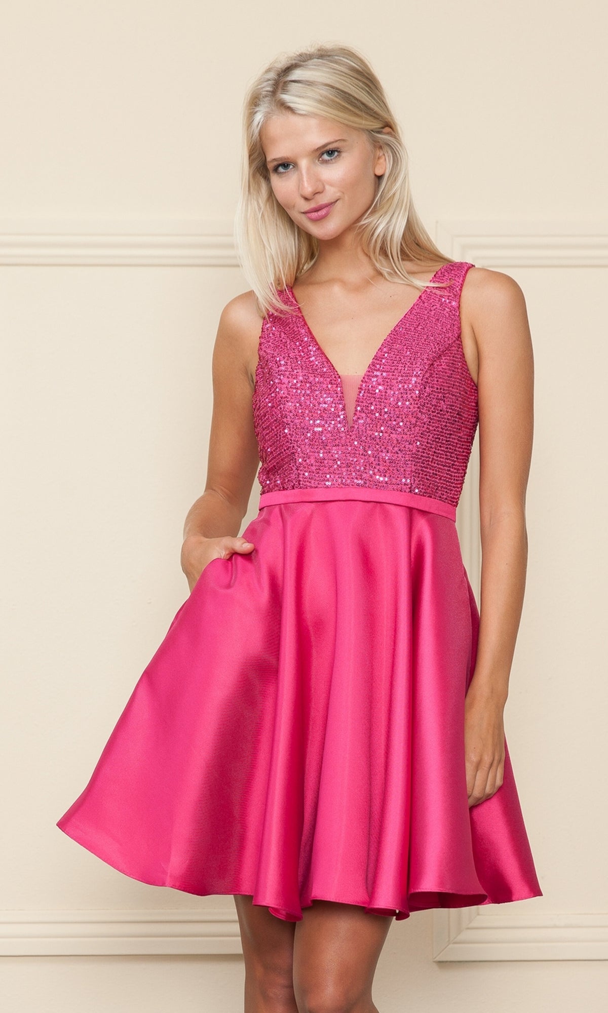 A woman wears the A-Line Short Homecoming Dress with Pockets - 8954, featuring a sleeveless pink sequin v-neck, embellished bodice, and flared skirt, standing against a plain beige background.